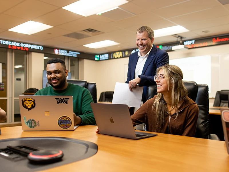 a professor stands behind two students on their laptops
