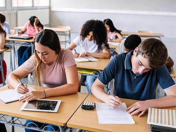 high school students writing at desks in a classroom, overseen by a teacher with a secondary education degree